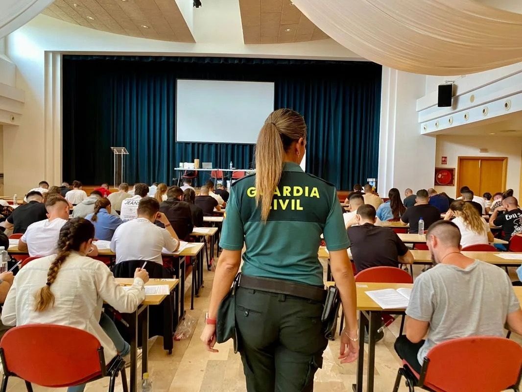 Salón de actos con opositores haciendo el examen en pupitres y una guardia civil de uniforme de espaldas en primer plano.