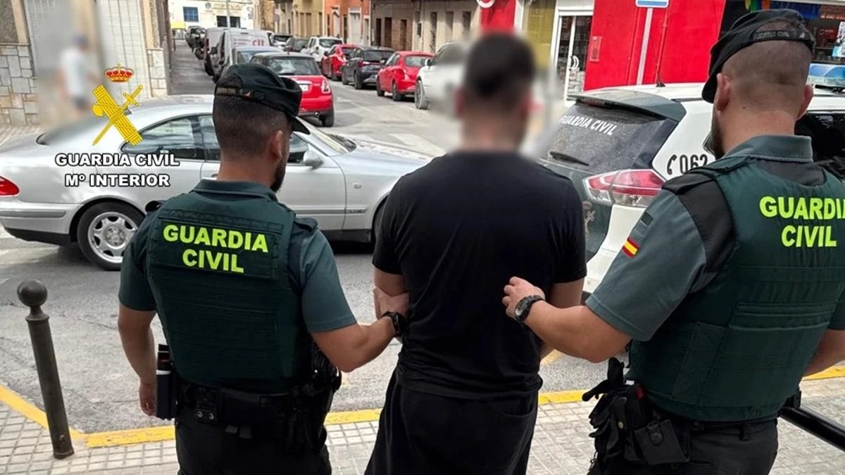 Dos guardias civiles de uniforme conducen esposado a un detenido junto a un coche patrulla en la calle.