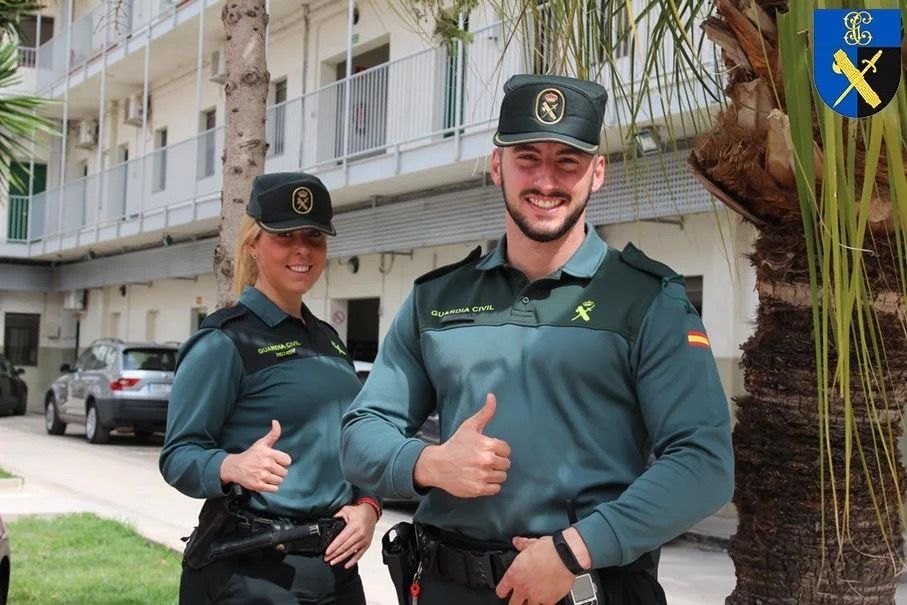Dos guardias civiles de uniforme, una mujer y un hombre, sonríen con el pulgar hacia arriba en el patio de un acuartelamiento.