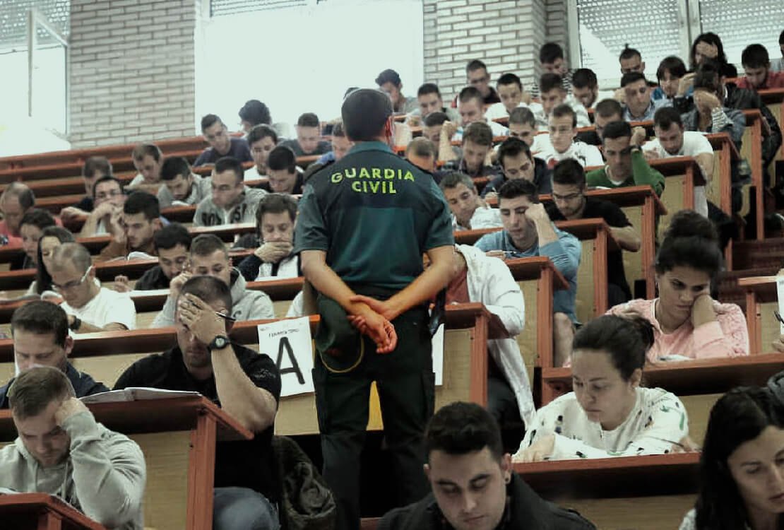 Aula llena de opositores haciendo un examen en pupitres individuales, con un guardia civil de uniforme vigilando entre las filas.